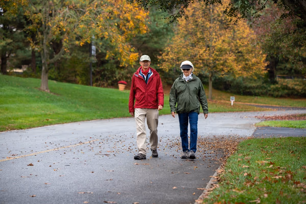Two older adults in jackets walking side-by-side on a tree-lined road with autumn leaves on the grass and pavement.