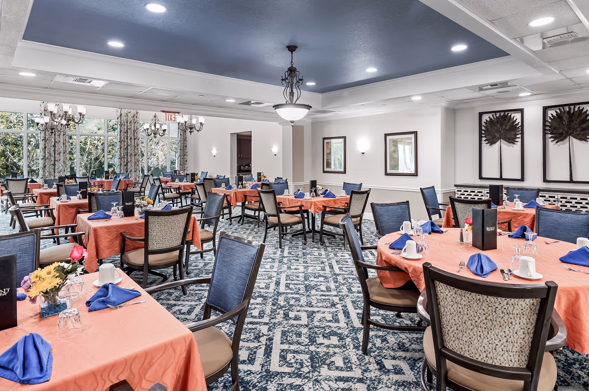 A spacious dining room with multiple round and rectangular tables covered with coral-colored tablecloths and set with blue napkins, white cups, glasses, and silverware. The room features patterned carpet, large windows with floral curtains, framed artwork on the walls, and ceiling lights including chandeliers.