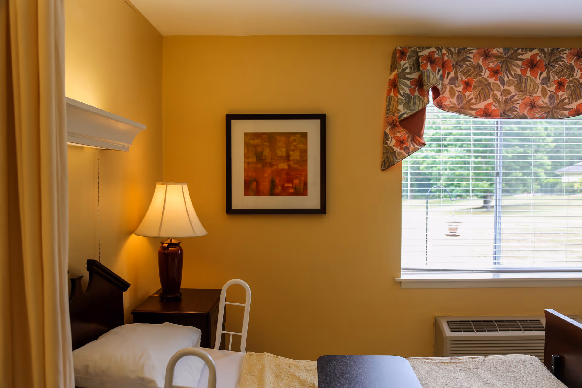 A cozy bedroom in a senior living facility with a single bed featuring white pillows and a beige blanket. Next to the bed is a wooden nightstand with a lamp that has a beige shade. Above the nightstand hangs a framed abstract painting. A window with floral-patterned valance allows natural light to brighten the room, and an air conditioning unit is installed below the window.