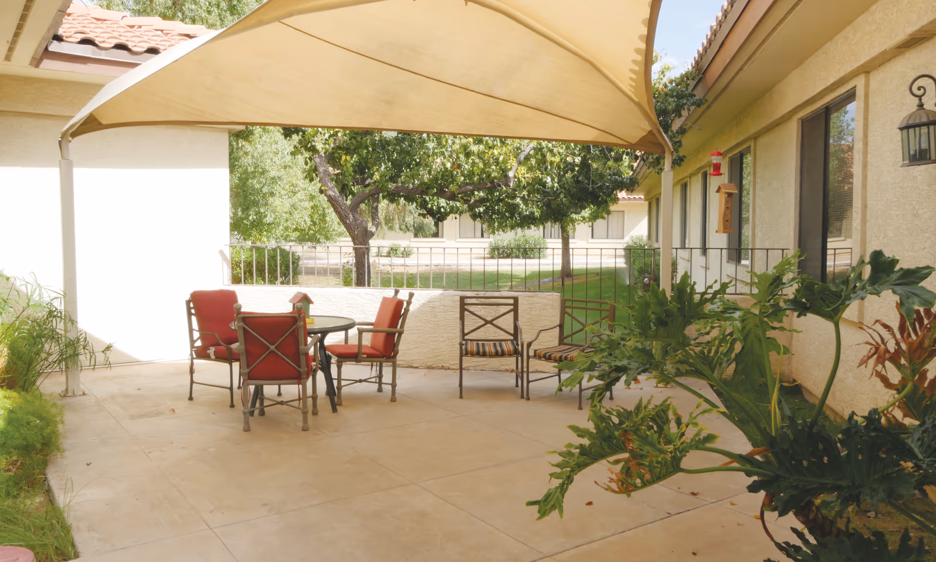 Outdoor patio area with a beige canopy overhead, four metal chairs with red cushions around a round glass table, two additional metal chairs with striped cushions, green plants on the right side, and trees and grass visible beyond a low wall and railing.