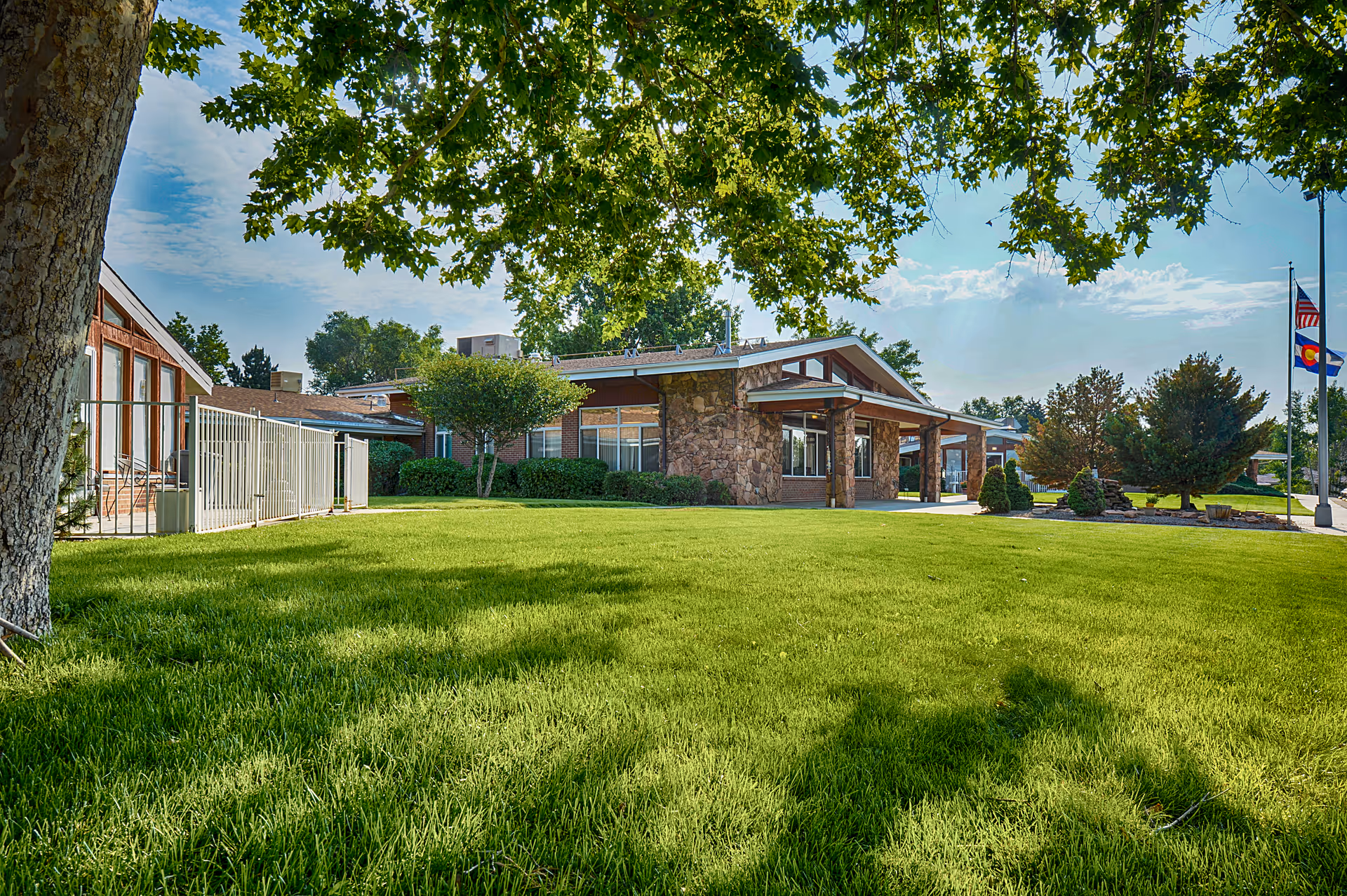 Exterior view of Harmony Pointe Nursing Center showing a single-story building with stone and brick facade, surrounded by a well-maintained green lawn and trees under a partly cloudy sky. Two flagpoles with flags are visible on the right side.