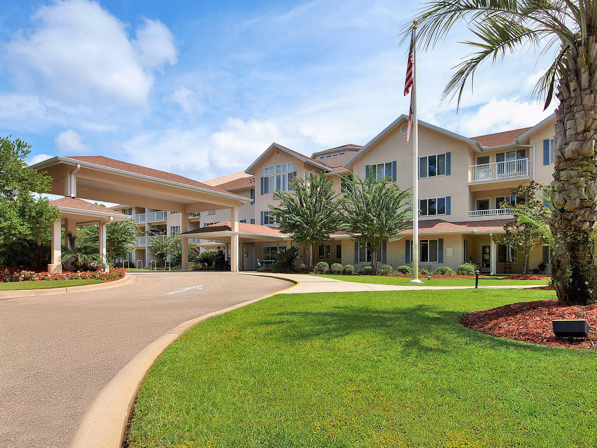Three-story senior living building with a covered drop-off entrance, flagpole, palm trees and manicured lawn under a blue sky.