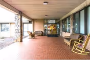 Covered outdoor patio area with brick flooring, rocking chairs, and benches along the sides. The entrance to a building with glass doors and windows is visible at the far end.