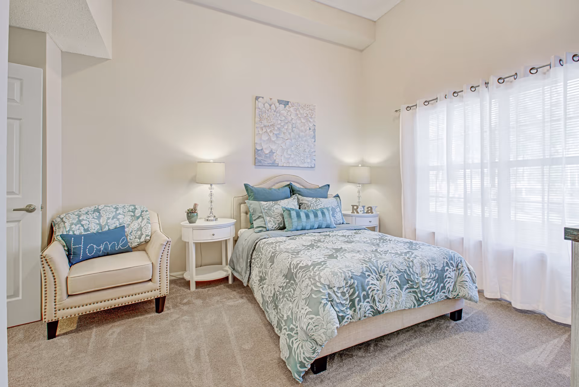 A bright bedroom with a bed dressed in blue and white floral bedding, two white nightstands with lamps on each side, a beige armchair with a blue pillow that says 'Home' and a matching throw, and a large window with sheer white curtains letting in natural light. A floral painting hangs above the bed.