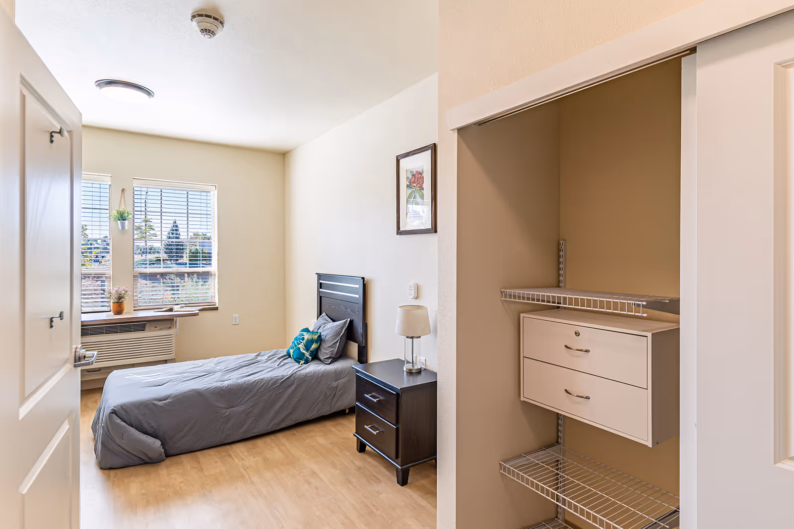Small, well-lit bedroom with a single bed, nightstand, window, and an open closet with wire shelving and drawers.