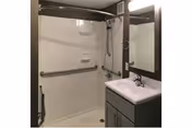 A clean, accessible bathroom featuring a walk-in shower with a grab bar, a handheld showerhead, and built-in soap holders. Next to the shower is a vanity with a sink, faucet, and a mirror above it. The bathroom has neutral-colored walls and a simple, functional design.