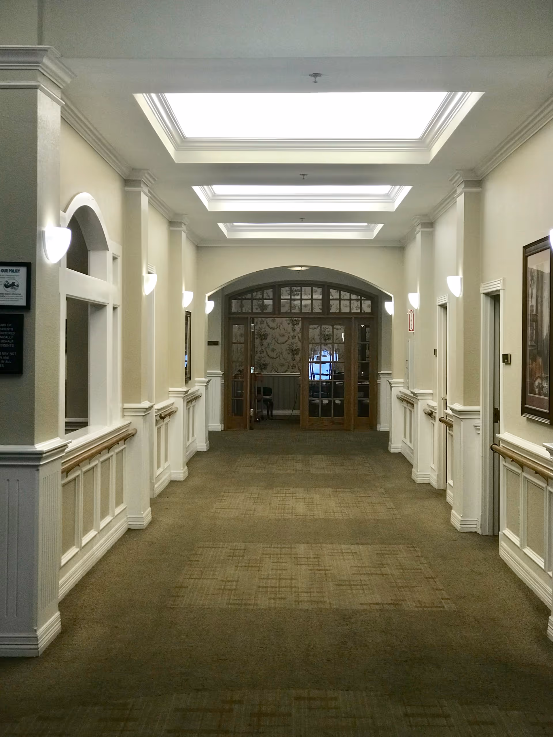 A well-lit hallway in a senior living facility with beige walls, white wainscoting, and carpeted floor. The ceiling features multiple rectangular skylights, and wall sconces provide additional lighting. At the end of the hallway, there are glass double doors with wooden frames leading to another room with patterned wallpaper.