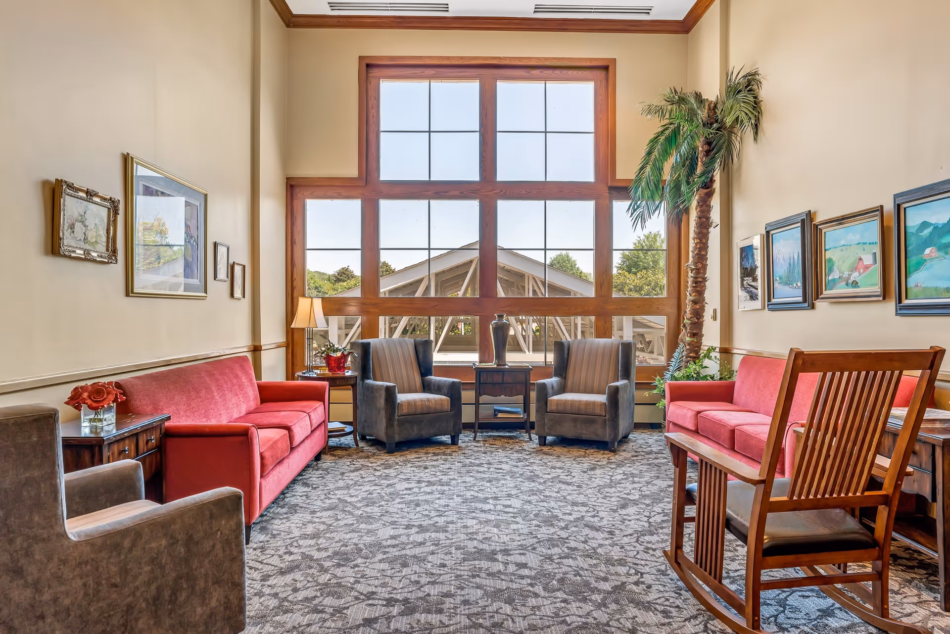 A cozy living room with two red sofas, two gray armchairs, a wooden rocking chair, and a large window with wooden frames letting in natural light. The walls are decorated with framed paintings and there is a tall artificial palm tree in the corner.