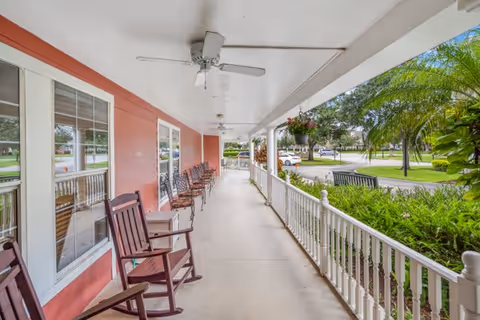 A covered outdoor porch area with several wooden rocking chairs lined up along a red exterior wall with windows. The porch has ceiling fans and white railings, overlooking a green landscaped area with trees and a parking lot in the background.