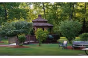 Wooded garden with a wooden gazebo, benches, a small bridge, and manicured lawn.