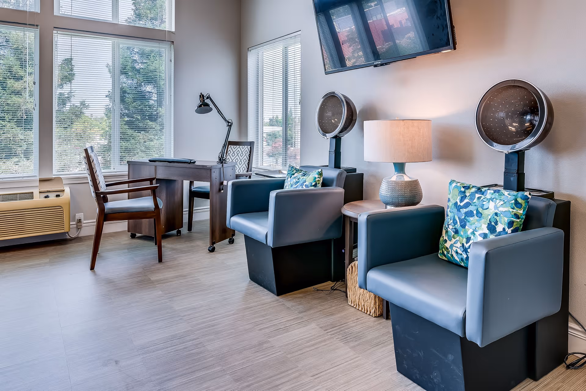 A bright room with large windows featuring two gray salon chairs with colorful cushions, two vintage hair dryers behind the chairs, a small round table with a lamp between the chairs, a wall-mounted TV above, and a wooden desk with two chairs near the windows.