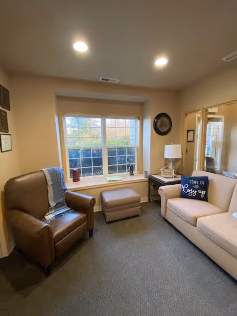 A cozy sitting area with a brown leather armchair draped with a blue and white blanket, a beige sofa with a decorative pillow that says 'Come in and cozy up,' a small ottoman, a side table with a lamp and decorative items, and a large window letting in natural light.