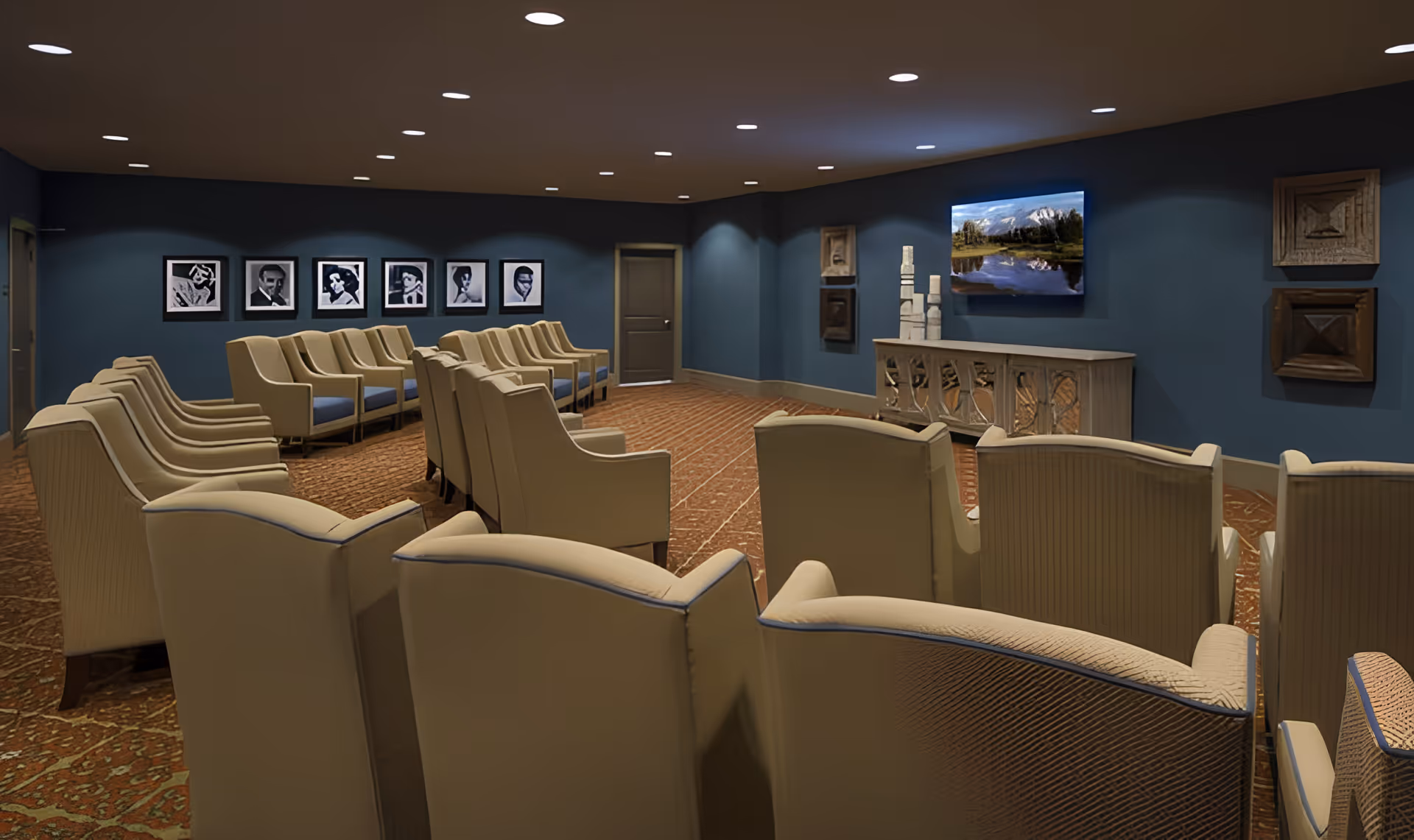 A carpeted communal media room with multiple rows of beige armchairs facing a wall-mounted TV and framed artwork.