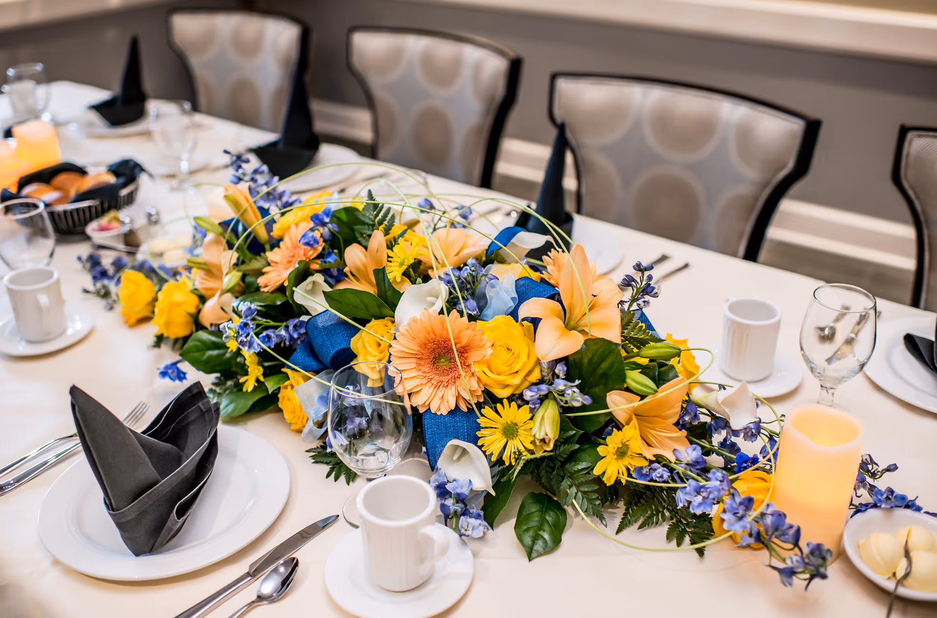A dining table set with white plates, black folded napkins, cups, glasses, and silverware. The centerpiece is a colorful floral arrangement with yellow, orange, and blue flowers, accompanied by a lit candle and a basket of bread rolls. Upholstered chairs with a circular pattern are arranged around the table.