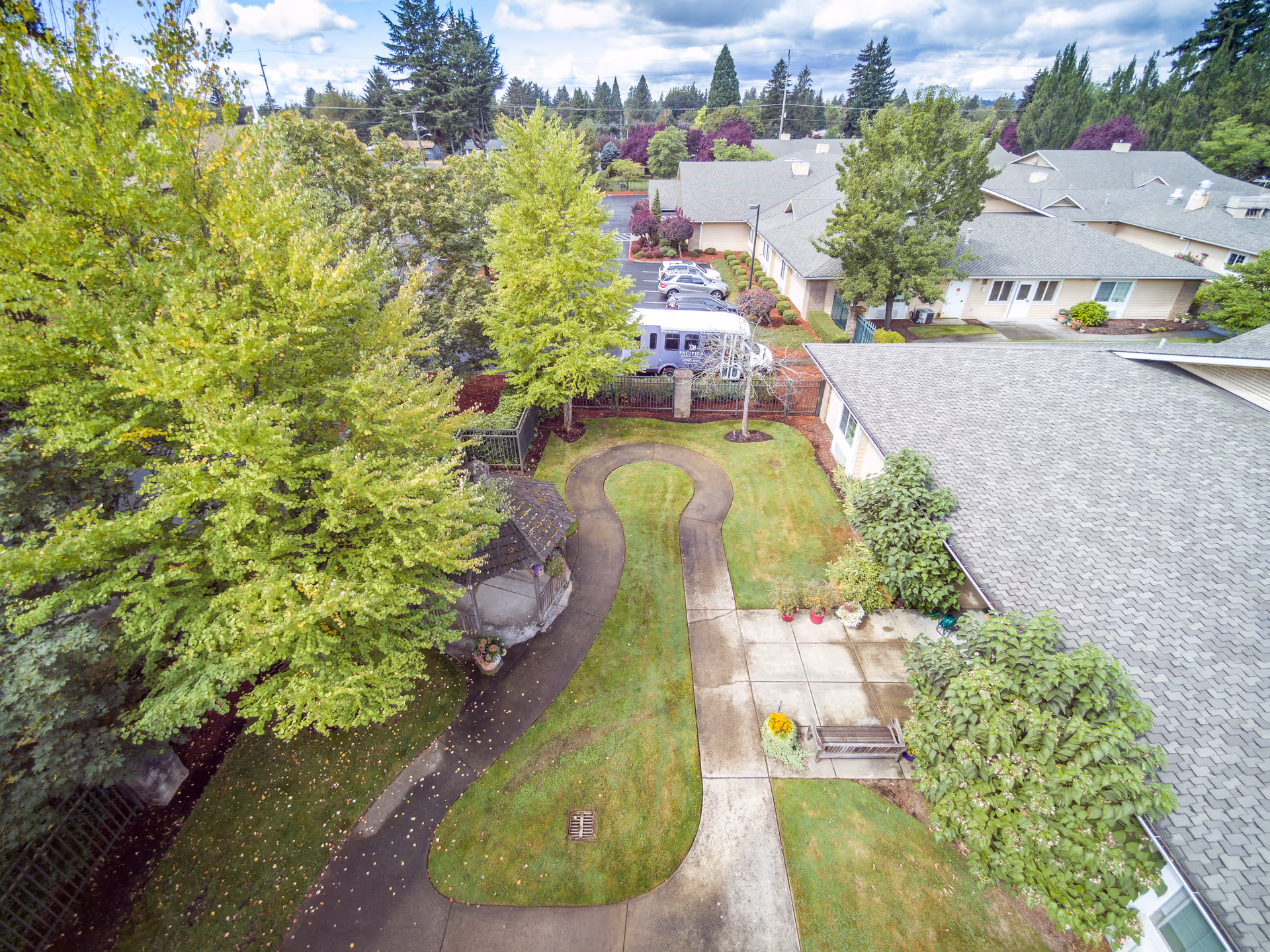Aerial view of a senior living facility's outdoor garden area with a winding paved pathway, green grass, trees, shrubs, and a small gazebo. Surrounding buildings with gray roofs and a parking lot with vehicles are visible in the background under a partly cloudy sky.