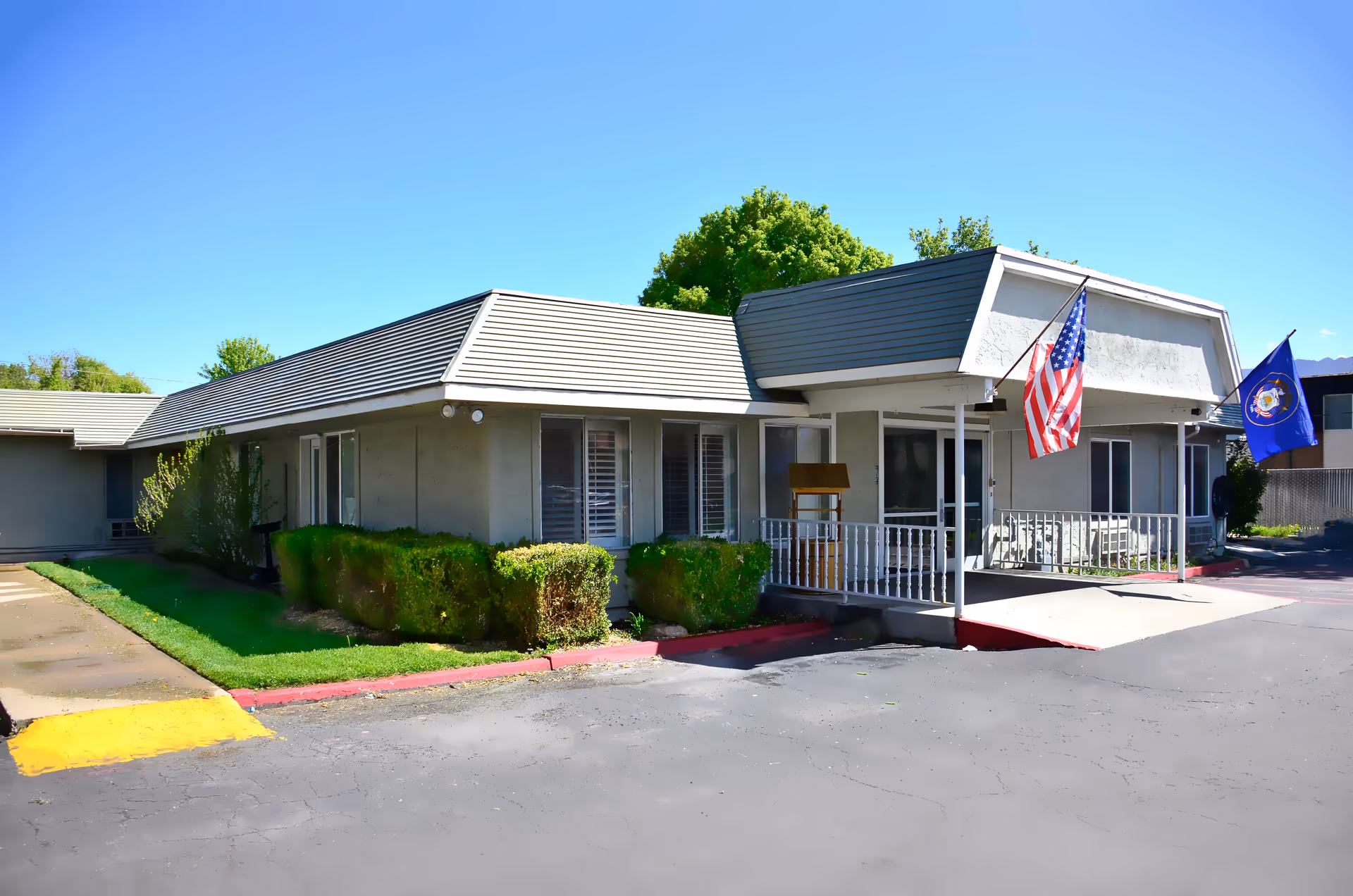 Single-story rehab and nursing facility entrance with an American flag, front porch, and low hedges under a clear sky.