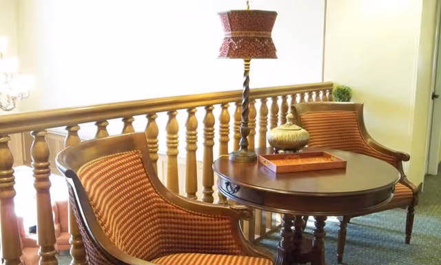 Two upholstered armchairs and a round wooden table with a lamp arranged beside a wooden balustrade in a seating area.