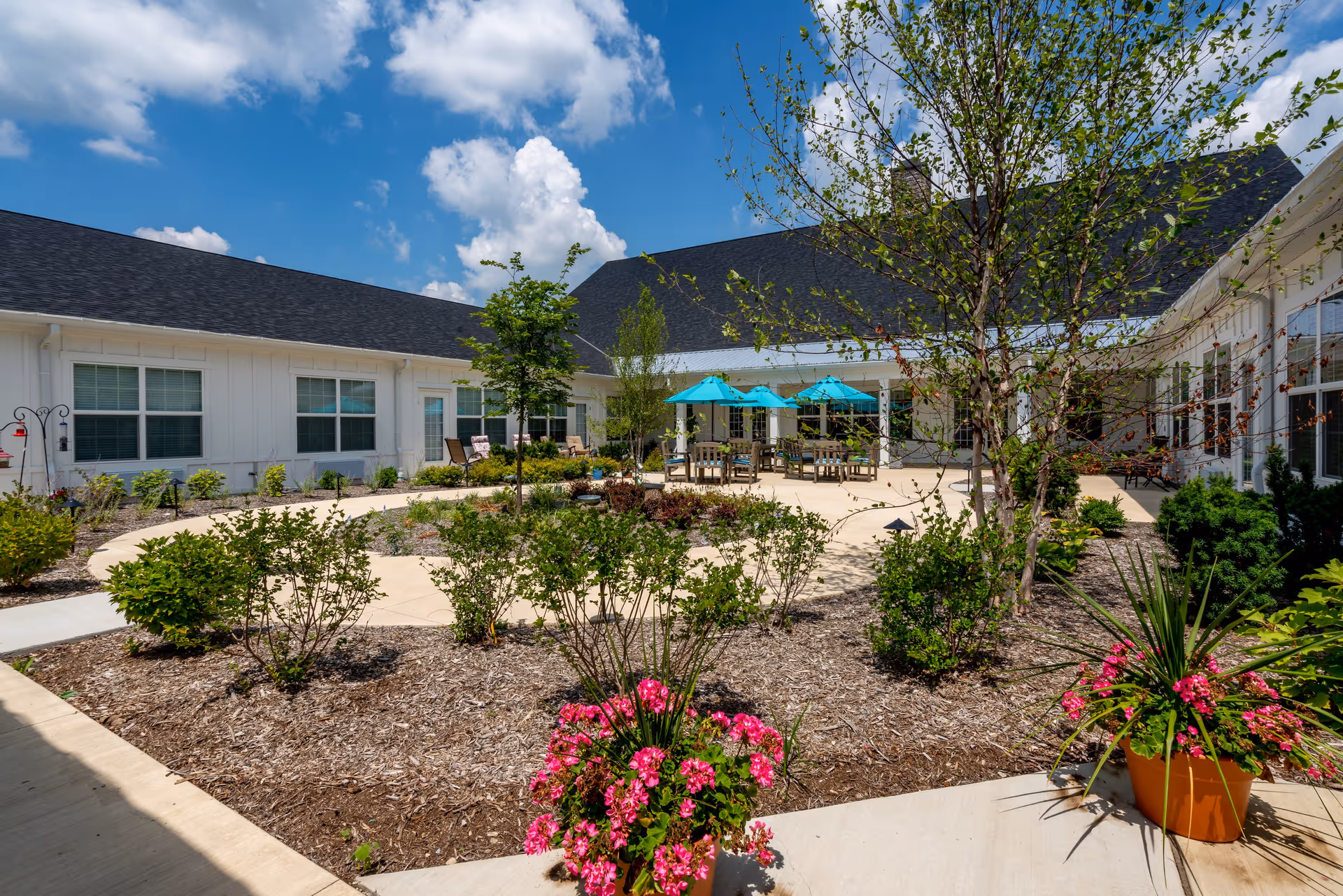 Outdoor courtyard area at Sugar Fork Crossing featuring a circular walkway, landscaped garden beds with various shrubs and flowers, several trees, and a seating area with tables and turquoise umbrellas under a bright blue sky with scattered clouds.