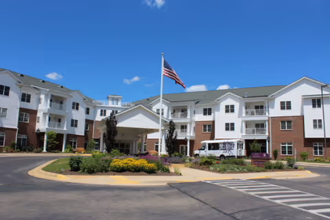 Front exterior of a multi-story senior living building with an entrance canopy, American flag, landscaped roundabout, and a parked van.
