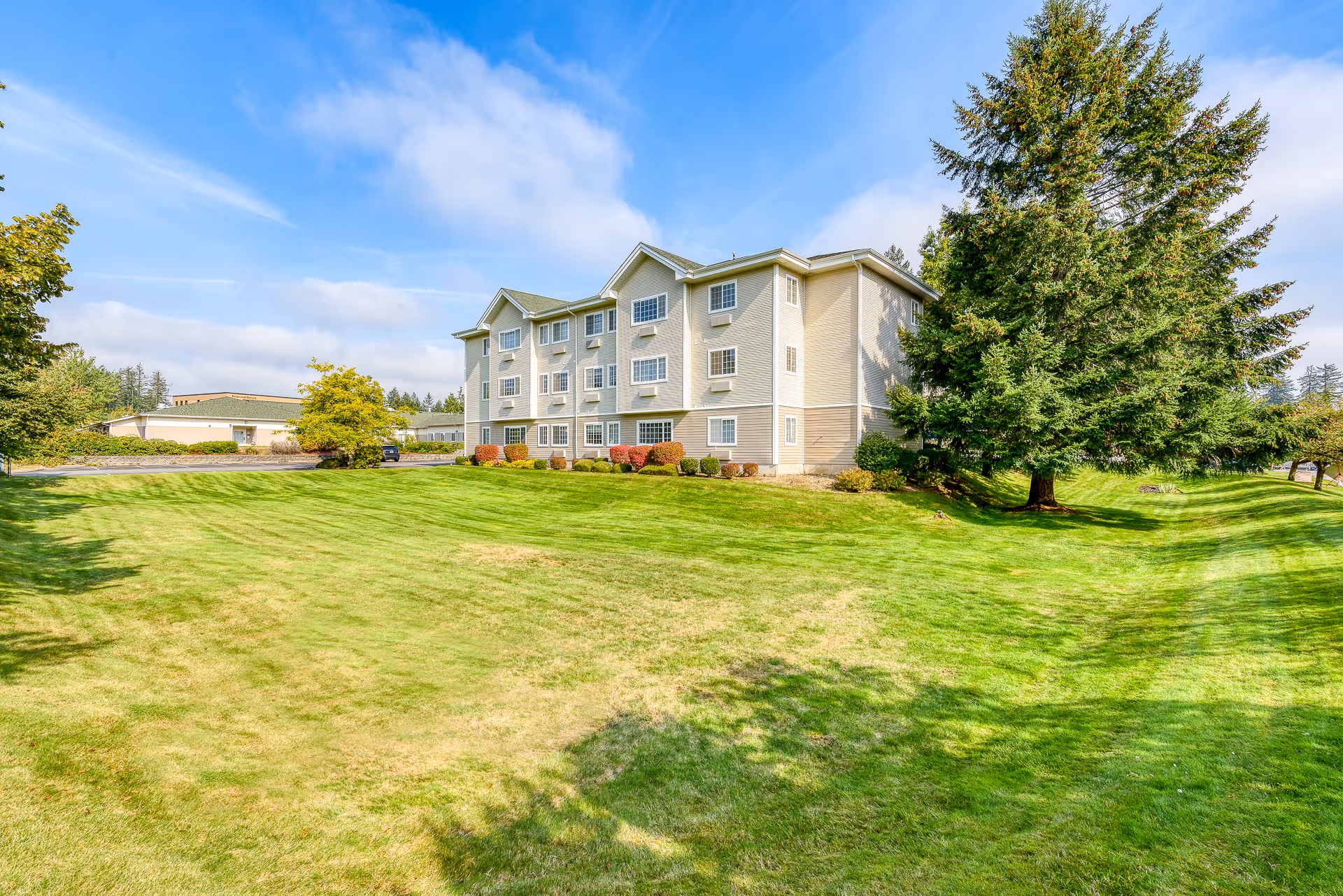 Exterior view of a three-story assisted living facility building with beige siding, surrounded by a well-maintained green lawn and trees under a partly cloudy blue sky.