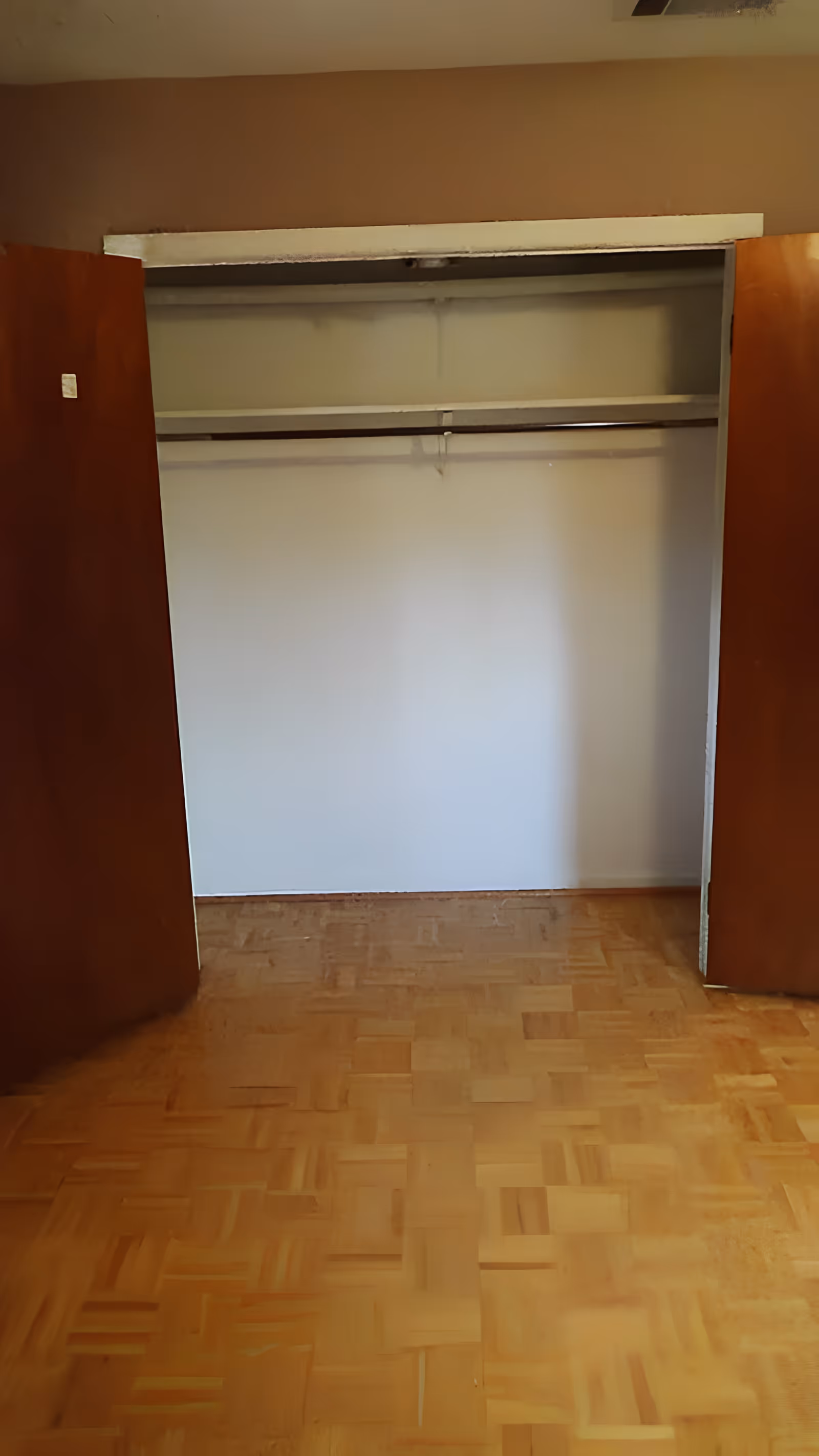 Empty open closet with wooden doors, a hanging rod and shelf above a parquet floor.