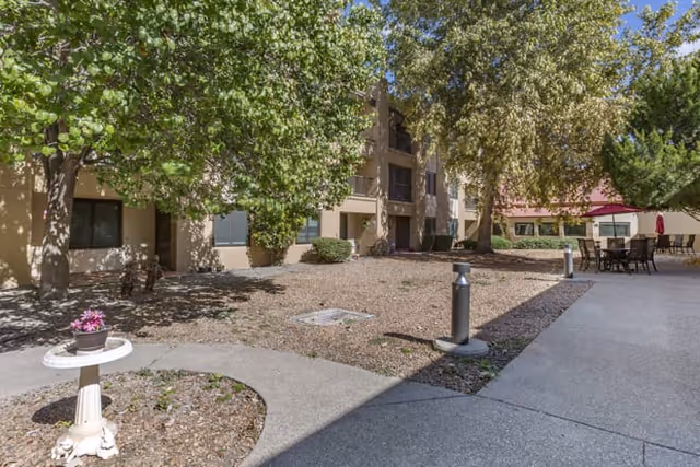 Courtyard with trees, paved walkways, patio seating and a beige multi-story residential building.