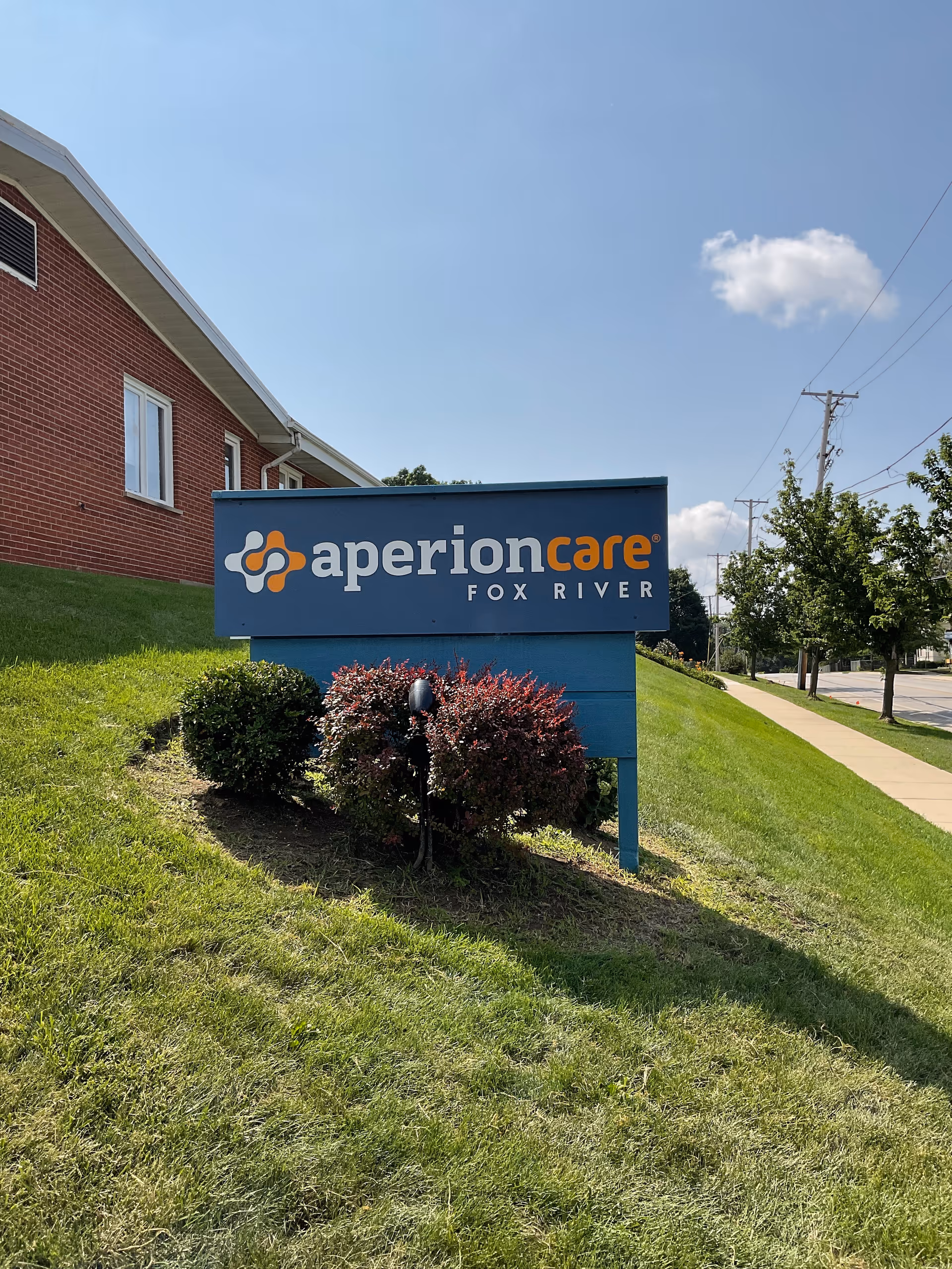 A blue sign reading 'aperioncare FOX RIVER' on a grassy slope in front of a brick building under a clear sky.