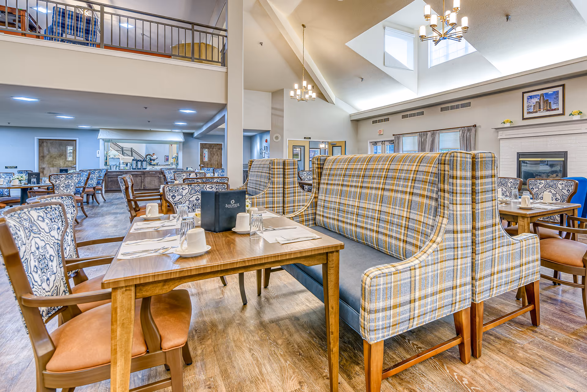 Bright dining room with plaid upholstered booths, wooden tables set for meals, chandeliers, and a fireplace.