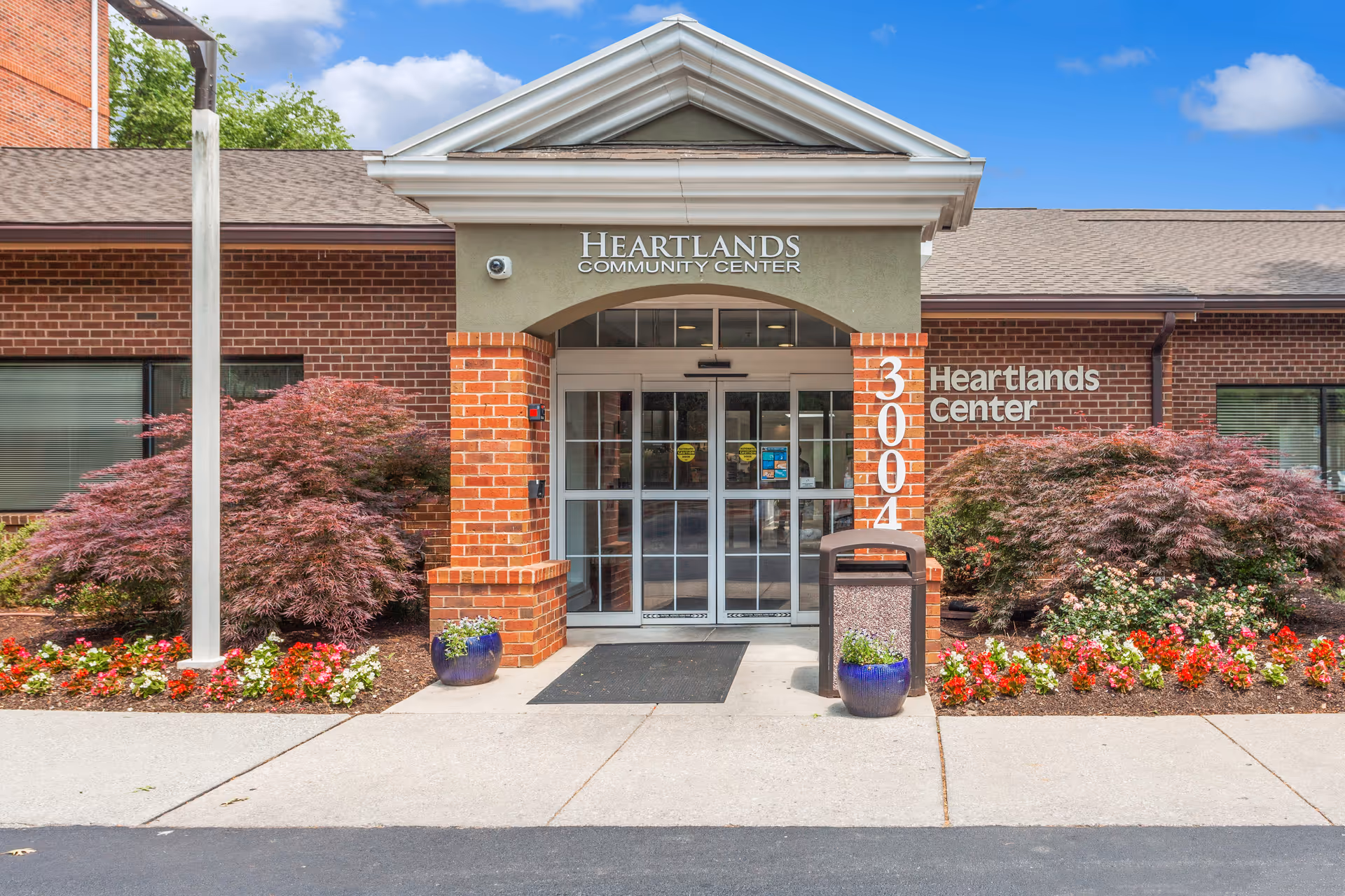 Front entrance of the Heartlands Community Center with brick facade, glass double doors, '3004' address column and landscaped flower beds.