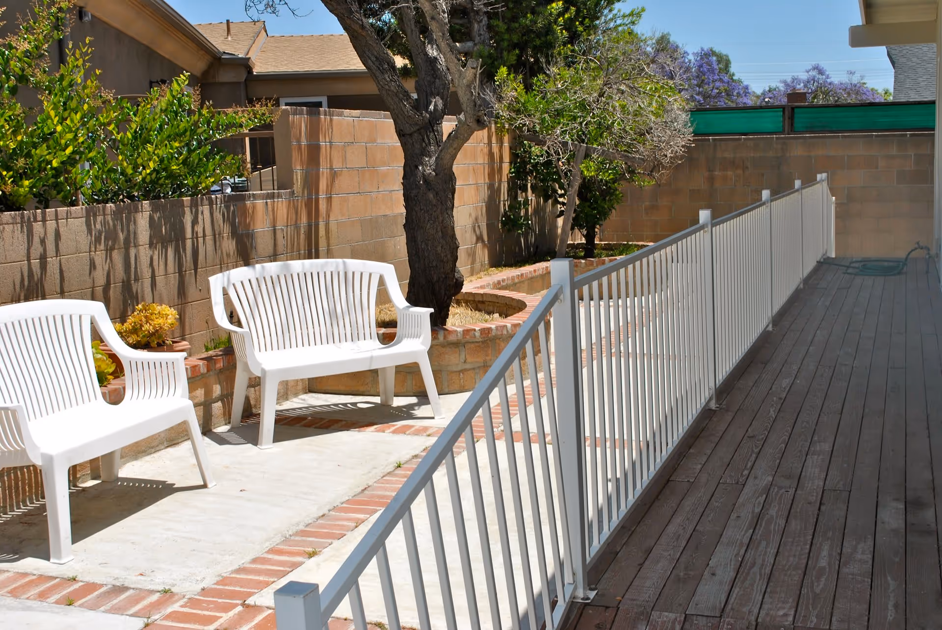 Outdoor patio area with two white plastic chairs and a white plastic bench placed on a concrete surface bordered by bricks. There is a tree planted in a raised brick planter along a brick wall, with some greenery and purple flowering trees visible in the background. A white metal railing separates the patio from a wooden deck area.