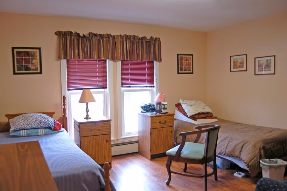 A simple bedroom with two single beds, each with pillows and blankets. Between the beds are two wooden nightstands, one with a lamp and the other with a phone and a cup. The room has two windows with red blinds and a striped valance. There are framed pictures on the beige walls and a wooden chair with a green cushion in front of one bed. The floor is wooden.