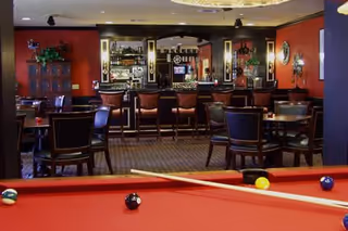 Interior view of a recreational lounge area featuring a red pool table in the foreground with several billiard balls and a cue stick on it. In the background, there are multiple tables and chairs arranged for seating, and a bar area with bar stools, shelves with bottles, and decorative lighting. The walls are painted red and adorned with framed pictures and plants.