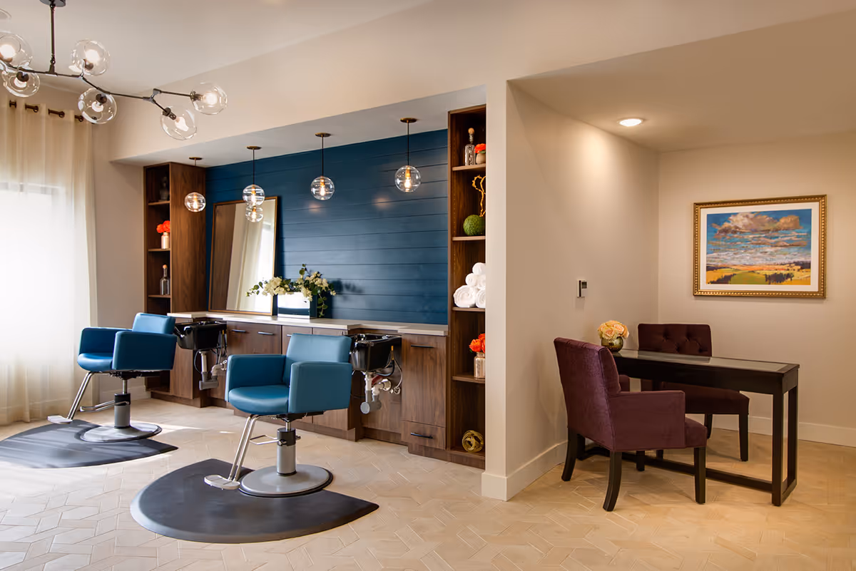 Interior view of a senior living facility's salon area featuring two blue salon chairs in front of a wooden counter with sinks and a large mirror. The wall behind the counter is painted blue with hanging globe pendant lights. To the right, there is a small seating area with a dark wooden table, two purple upholstered chairs, a vase with flowers, and a framed landscape painting on the wall.