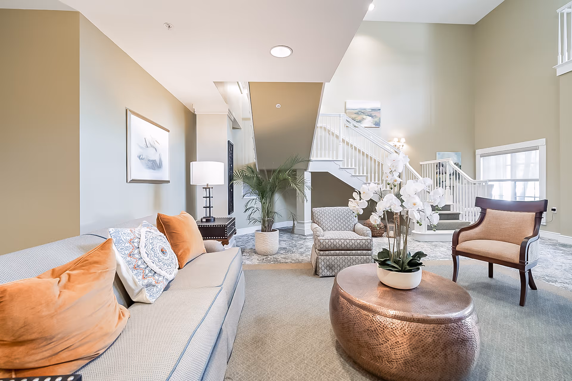 A bright and spacious living room area with a beige sofa adorned with orange and patterned pillows, a round copper coffee table with a white orchid plant, an upholstered armchair, and a wooden chair with beige cushions. In the background, there is a staircase with white railings and a potted plant near the wall. The walls are painted light beige and decorated with framed artwork.