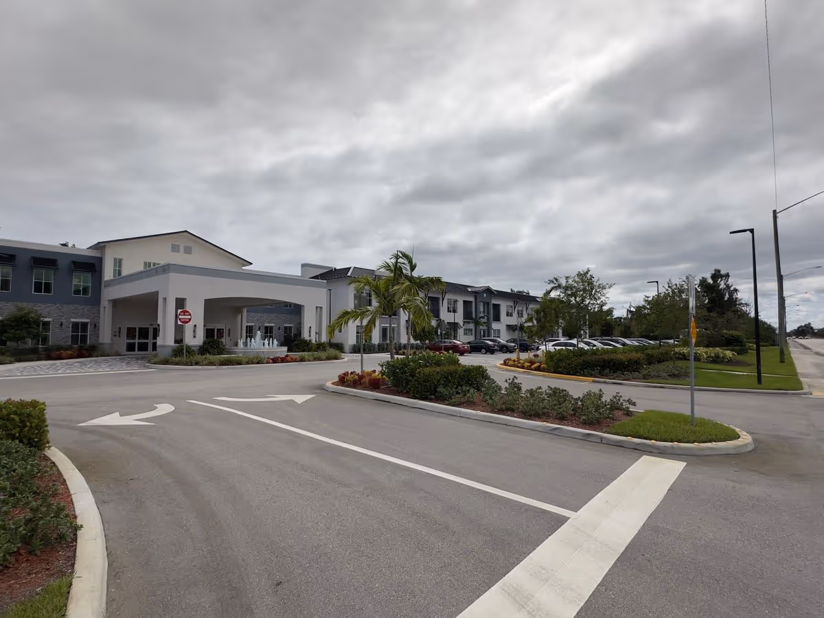 Entrance driveway and porte-cochère of a two-story senior living building with landscaped roundabout and parked cars under an overcast sky.