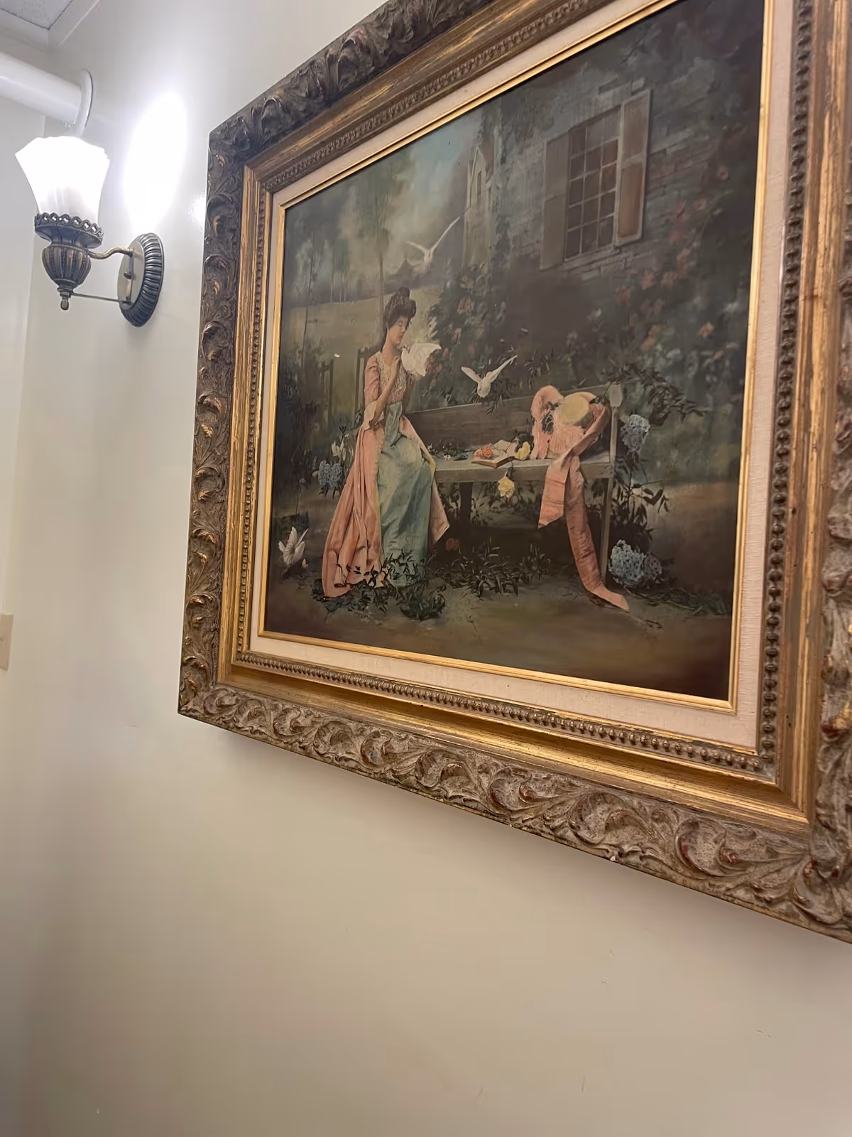 Ornate framed painting of a woman seated on a bench with doves, hanging on an interior wall next to a lit wall sconce.
