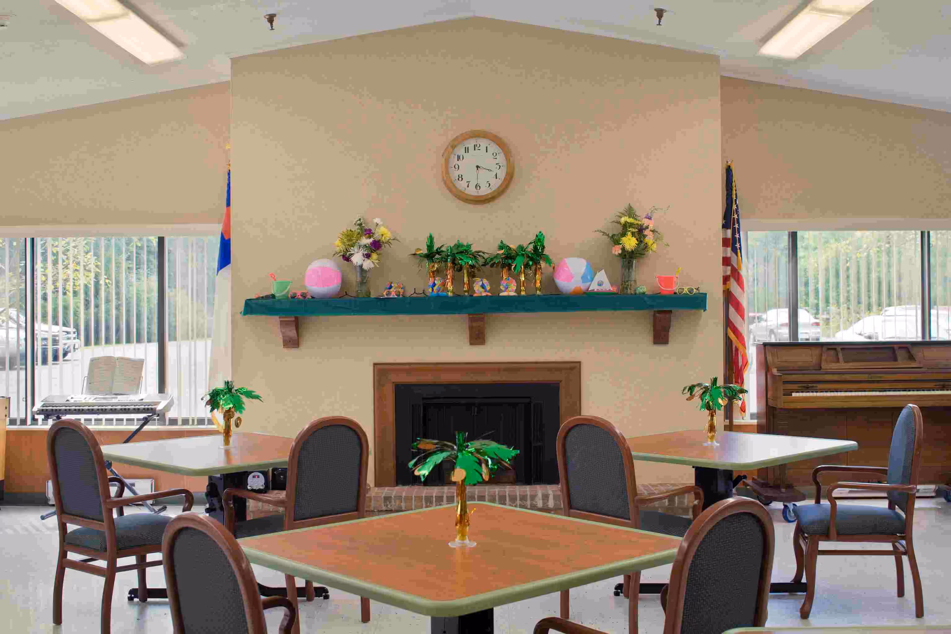 A common area in a senior living facility with four tables, each surrounded by chairs. The tables have small decorative palm tree centerpieces. A fireplace with a green mantel decorated with flowers and ornaments is centered on the far wall, with a clock above it. Large windows on either side let in natural light, and an upright piano is visible on the right side of the room. Two flags are positioned near the windows.