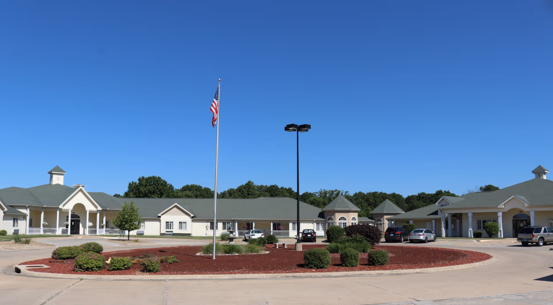 Front exterior of a single-story senior living complex with a circular driveway, flagpole, and parked cars under a clear blue sky.