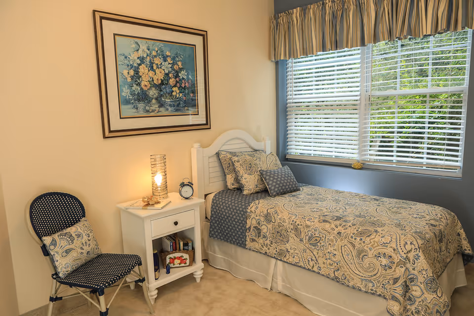 Single bedroom with a twin bed, patterned bedding, a white nightstand holding a lamp and alarm clock, a chair, and a large window with blinds.