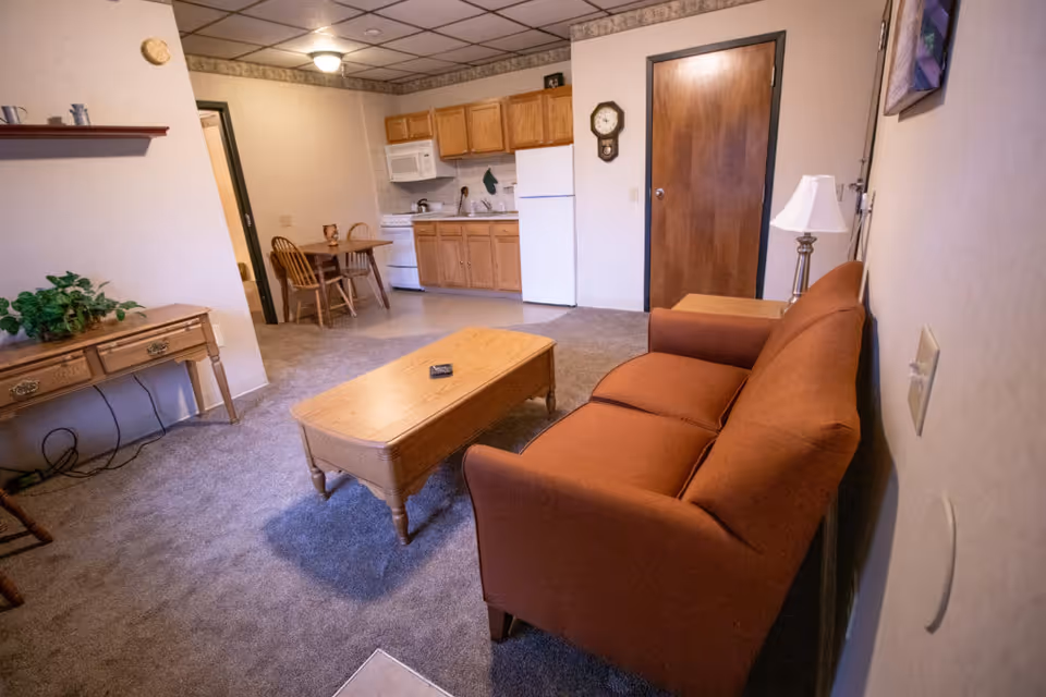 Interior view of a living area in a senior living facility showing a brown sofa, wooden coffee table, side table with a lamp, a small dining table with two chairs, and a kitchenette with wooden cabinets, a refrigerator, microwave, and stove. The room has carpeted flooring and beige walls with a wall clock and a framed picture.