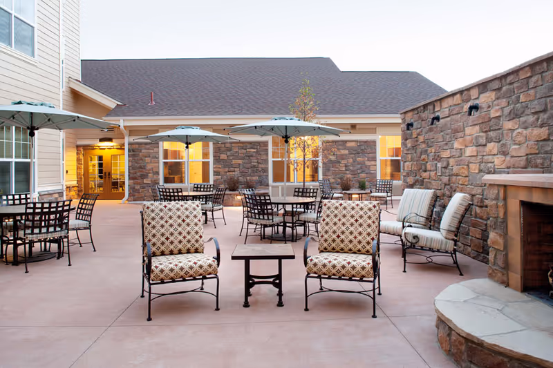 Outdoor patio area at MorningStar Assisted Living & Memory Care at Jordan with multiple tables and chairs, umbrellas, cushioned armchairs, and a stone fireplace against a stone wall. The building exterior has beige siding and stone accents with windows and a door leading inside.