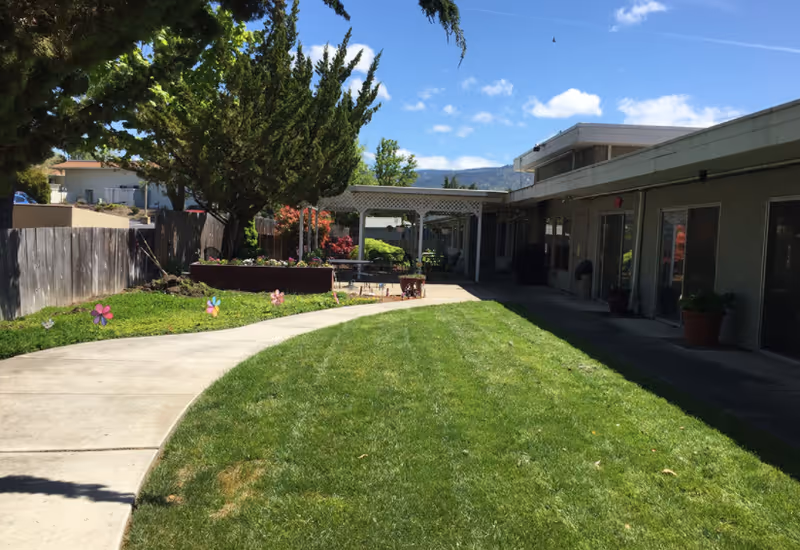 Outdoor area of Hearthstone Nursing & Rehabilitation Center featuring a curved concrete walkway, green lawn, potted plants, trees, and a covered patio area attached to the building under a blue sky with some clouds.