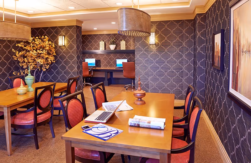 A well-lit interior room with wooden tables and red cushioned chairs arranged around them. On the tables are a laptop, papers, a rolled-up magazine, and decorative items. The walls have a patterned wallpaper with framed artwork and wall sconces. In the background, there is a small workspace with two computer monitors and red office chairs. A large decorative plant is also visible.