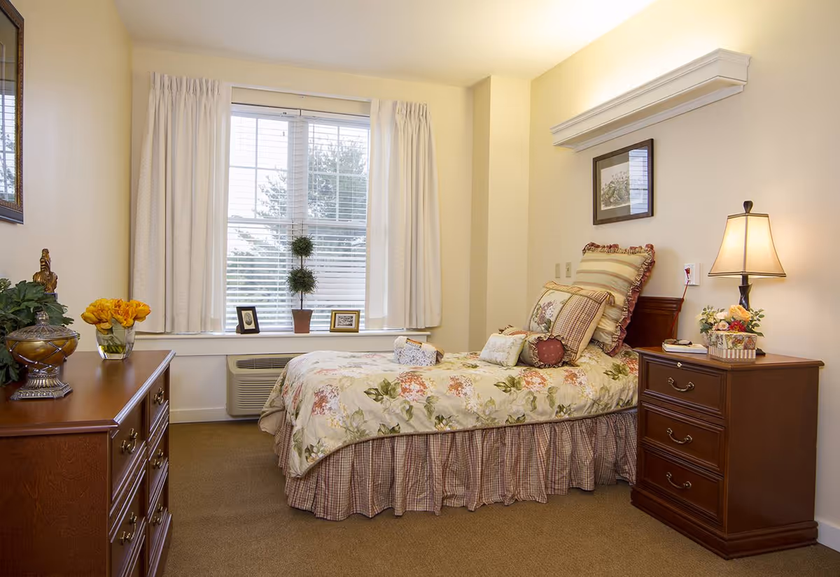 A cozy bedroom with a single bed covered in floral bedding and multiple pillows. There is a wooden nightstand with a lamp and a small flower arrangement on the right side of the bed. On the left side of the room, there is a wooden dresser with decorative items including a vase with yellow flowers. A window with white curtains lets in natural light, and a framed picture hangs on the wall above the bed.