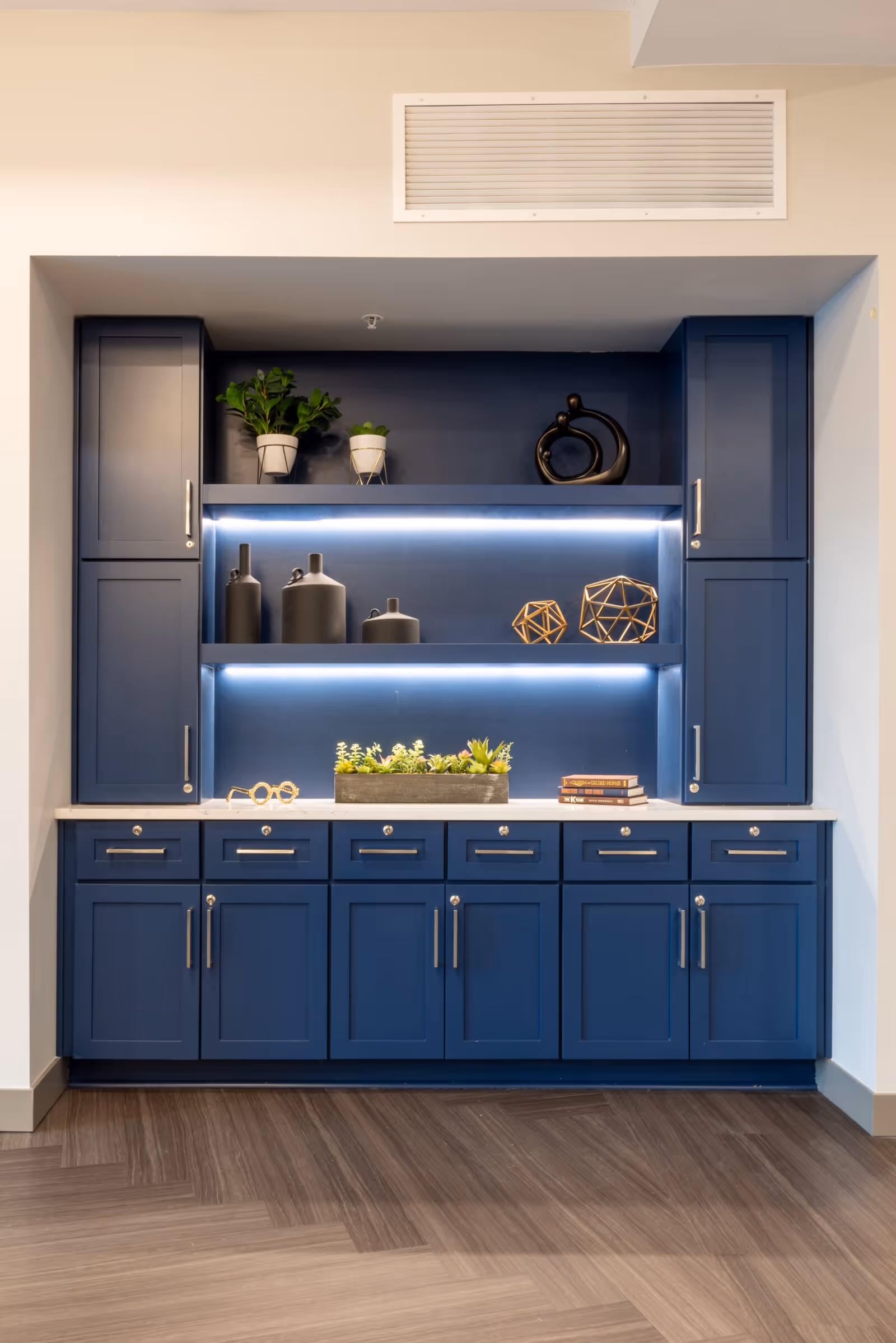 Built-in blue cabinetry with multiple drawers and cupboards, featuring two open shelves with decorative items including potted plants, black vases, geometric sculptures, and a small stack of books. The cabinetry is set against a beige wall with a vent above and a wood-patterned floor below.