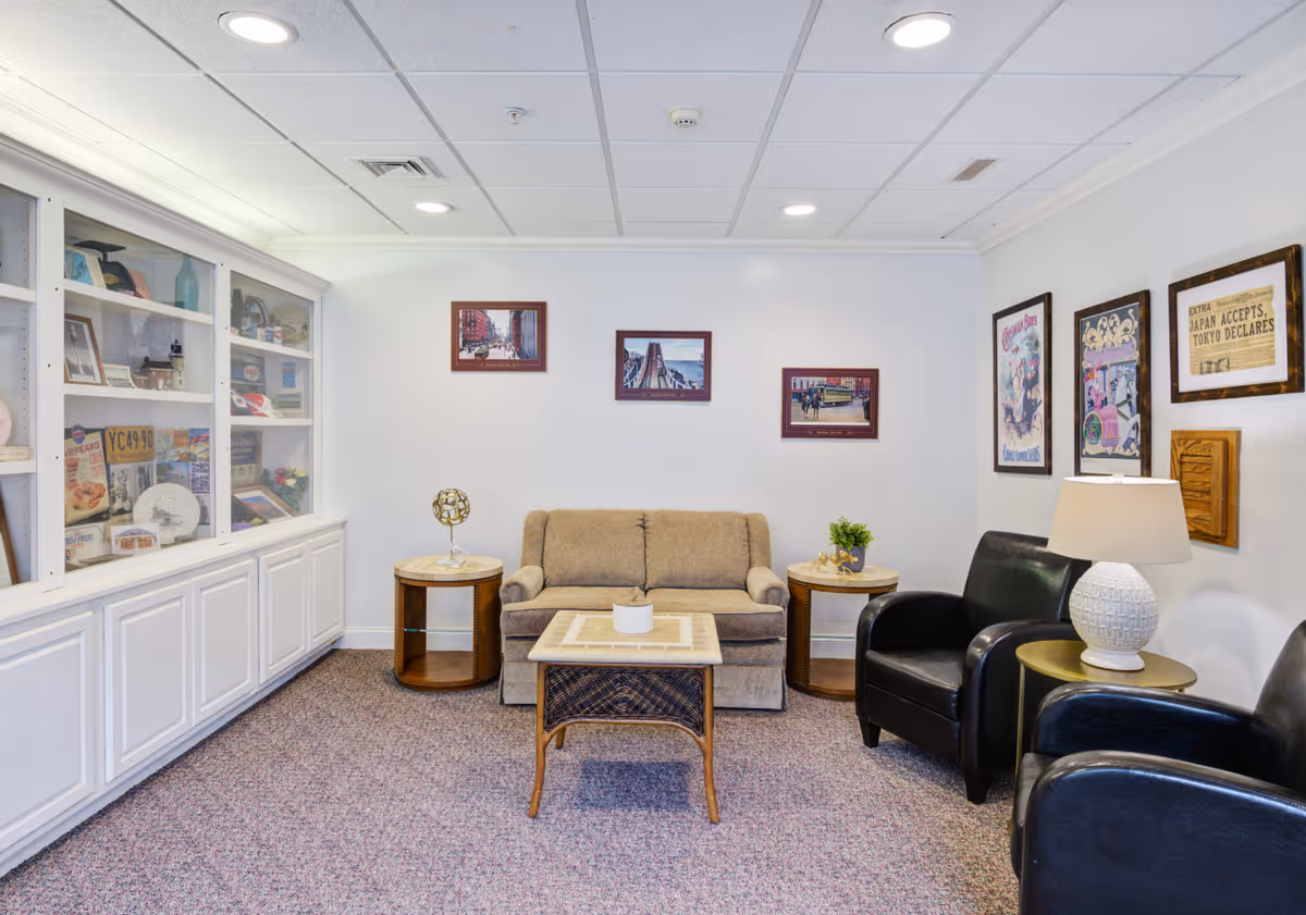 A cozy sitting area in a senior living community featuring a beige loveseat, two black armchairs, a wicker coffee table, and two round side tables with decorative items. The walls are adorned with framed pictures and posters, and a built-in white cabinet with glass doors displays various memorabilia. The room has a carpeted floor and recessed ceiling lights.
