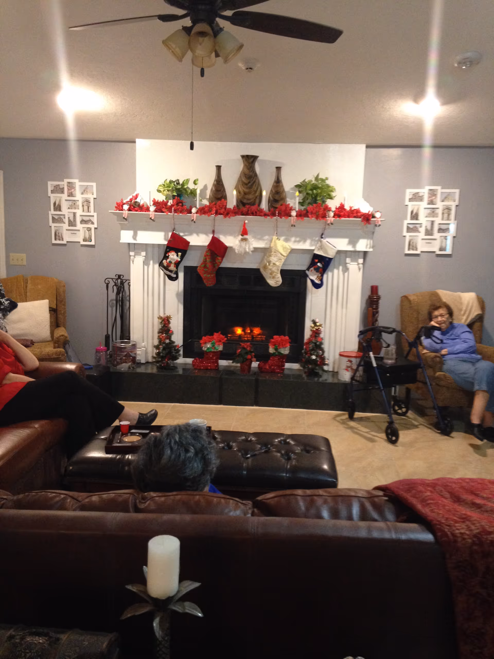 A cozy living room with a lit fireplace decorated for Christmas with stockings and red garlands. There are three people seated on couches and chairs around a central ottoman. The room has a ceiling fan with lights, framed pictures on the walls, and a walker near one of the seated individuals.