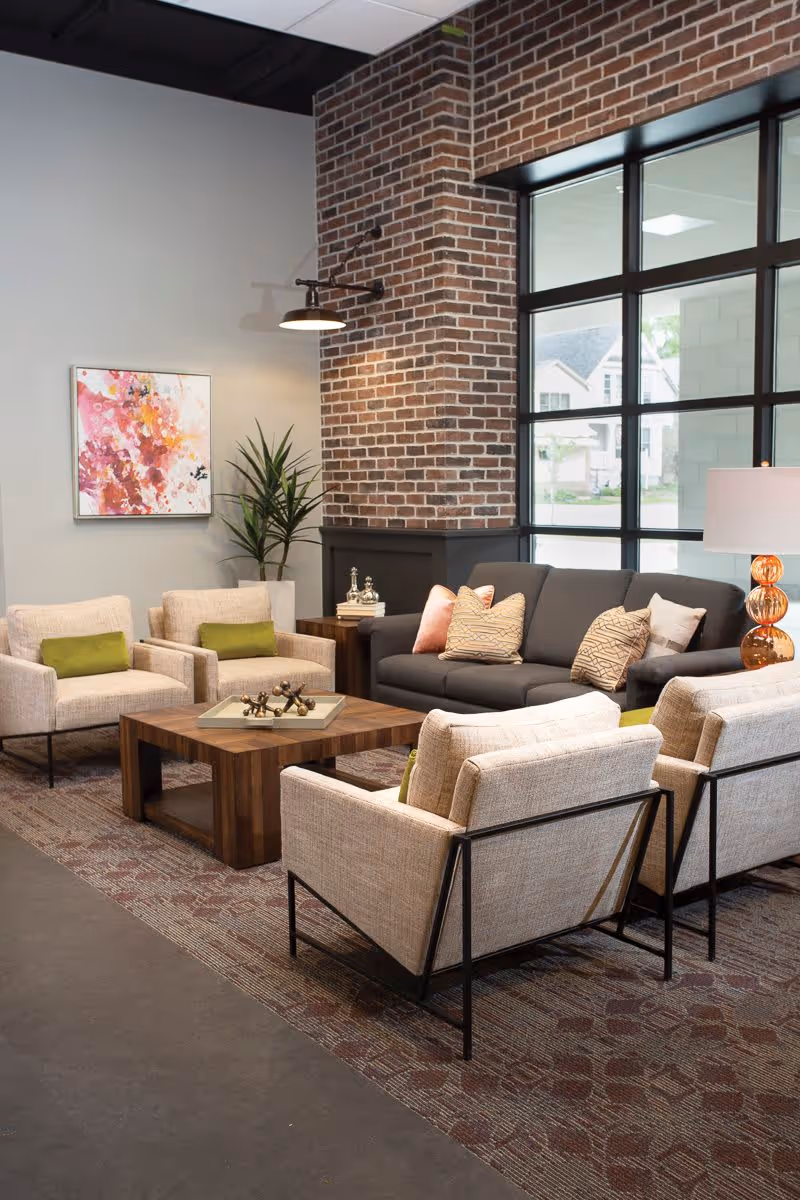 A cozy living room area with a dark gray sofa adorned with patterned and solid pillows, surrounded by four beige armchairs with green cushions. A wooden coffee table with decorative items sits in the center on a patterned rug. The room features a brick accent wall with a mounted light fixture, a large window with black framing, a potted plant, a side table with books and decorative objects, and a lamp with an orange glass base.
