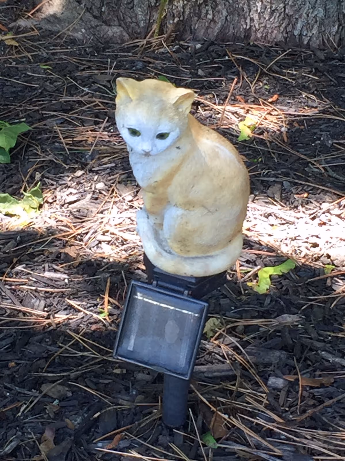 A small cat statue perched on a garden solar light in mulch beneath a tree.