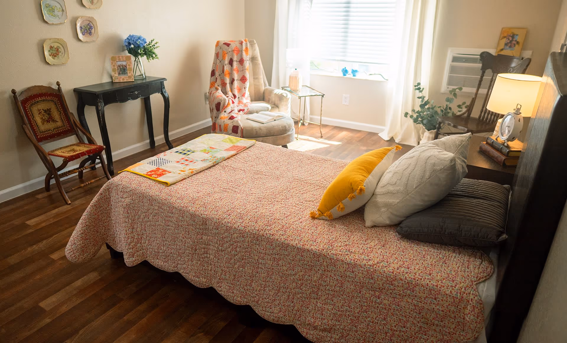 A cozy bedroom in a senior living facility with a bed covered in a floral quilt and multiple pillows. The room features wooden flooring, a cushioned armchair with a colorful throw blanket, a small side table with a lamp, a black console table with a vase of blue flowers and a framed photo, and decorative plates hanging on the wall. There is also a wooden chair and an air conditioning unit under the window with white curtains.